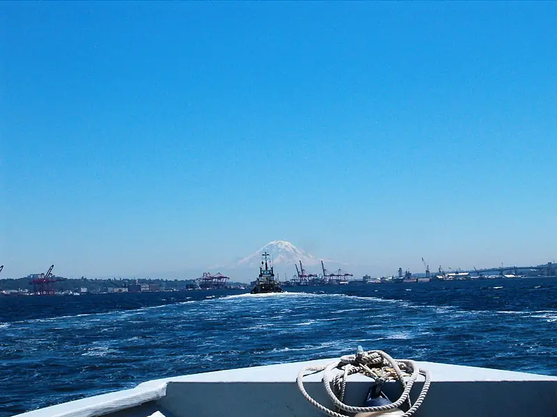 Tugboat heading away from frame towards Mount Rainier and large cranes visible in the distance.